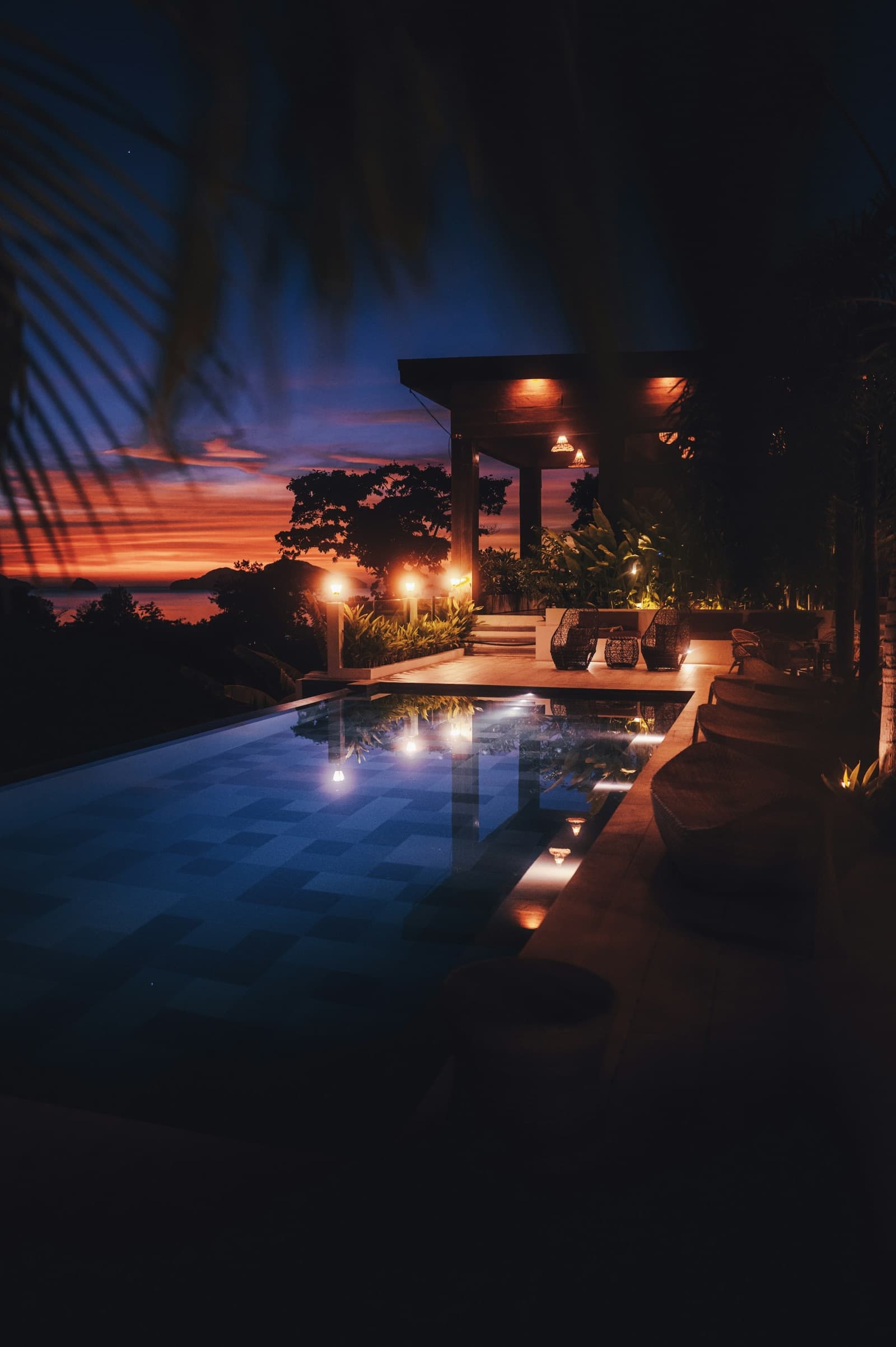Terrasse einer privaten Villa mit beleuchtetem Pool in der Abenddämmerung, eingerahmt von Palmblättern.